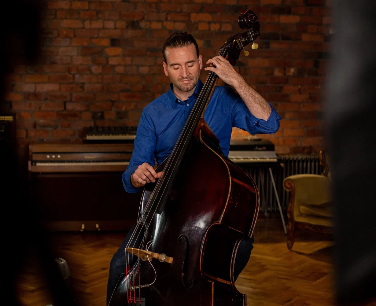 Man playing a double bass in a room with brick walls and musical instruments.
