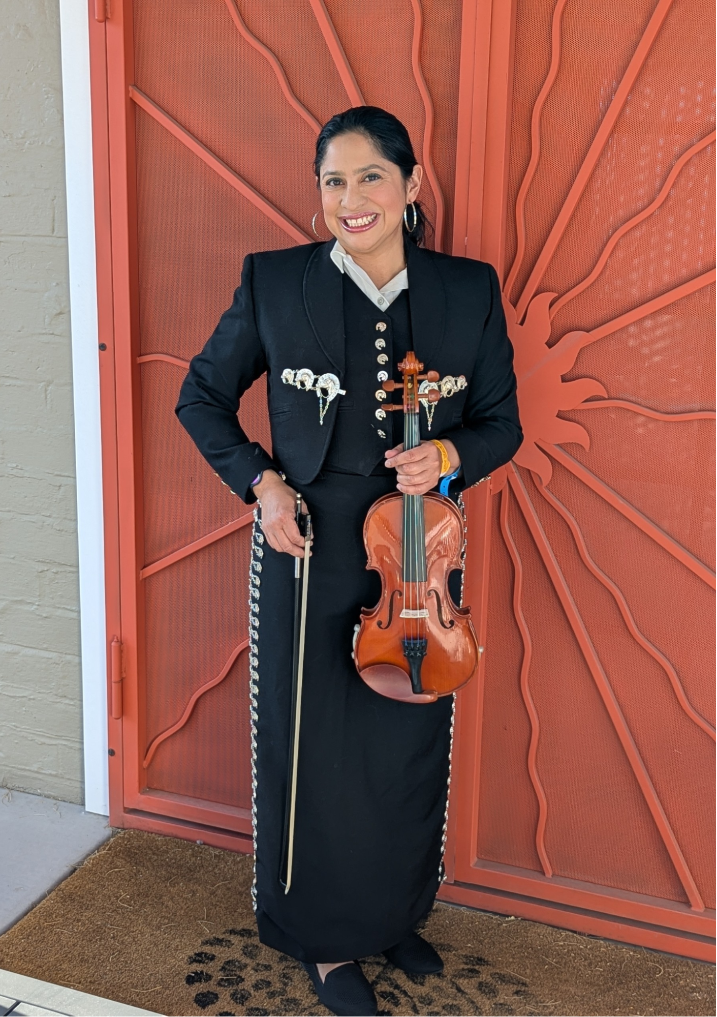 Person in traditional mariachi outfit holding a violin in front of a decorative red door.